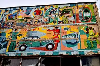 A large, colorful, and partially crumbling Soviet-era mosaic on a brick wall. The mosaic depicts industrial scenes with workers, tools, and a vintage blue car.