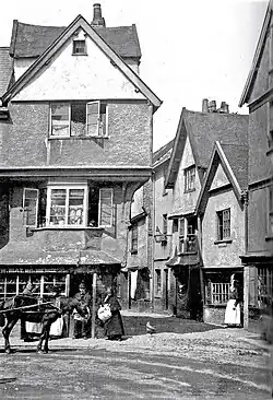 Image of the front of the house whilst at 16 Edumund Street, with a horse and carriage outside, and one man and three women posed outside, and a sign which reads W May Boot and Shoe Maker.