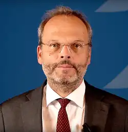 European man in a suit, red tie, and glasses in front of a dark blue background
