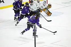 A purple-clad Minnesota Frost player shoots a puck towards three white-clad Boston Fleet players protecting the goal. Three other Minnesota players rush towards the puck.