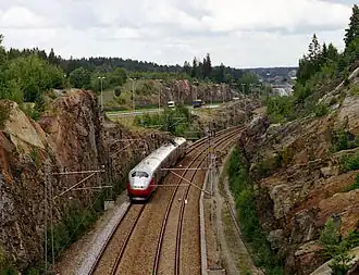 A four-car multiple unit running along a double-track line between two cliffs; in the background is a motorway.