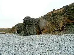 a cliff of folded grey and brown rocks, looming over a beach of grey cobbles