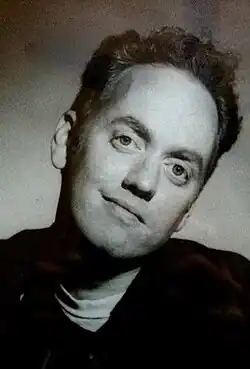 A black-and-white image of a man with short dark hair and a sweater with a white shirt underneath. He is looking toward the camera.