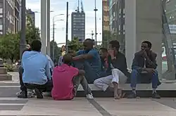 A group of African men sitting or squatting on a low bench next to a glass wall in a large city square. In the rear can be seen a street with a tall rectilinear skyscraper