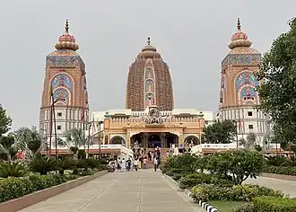 The image showcases the grand entrance to the **Agroha Dham Hindu temple**, with three towering, ornately decorated structures rising prominently against an overcast sky. The central structure is the tallest, with a distinctive brownish-red facade adorned with symmetrically placed windows and carved niches that add depth and texture. Its dome has a rounded, layered design crowned with a golden pinnacle at the top. Flanking the central tower are two slightly shorter towers, each adorned with vibrant colors and intricate artwork. Both towers have sky-blue panels accented with floral patterns and traditional Hindu motifs in hues of pink, gold, and red. The top of each of these side towers is capped with lotus-like structures, topped by golden finials. At the main entrance of the temple, a statue depicting a divine chariot pulled by horses and guided by warriors or deities stands prominently, drawing attention. Below the chariot, a large archway leads into the temple, with detailed architectural elements on either side. The steps leading up to the entrance are wide and surrounded by well-maintained greenery, with various plants, shrubs, and flowering bushes lining the walkway. The scene is bustling with visitors, some ascending the stairs towards the temple, while others are seen standing or moving around in groups. The pathway leading up to the temple is clean and well-kept, bordered by a trimmed lawn and decorative plants, contributing to the peaceful and reverent atmosphere.