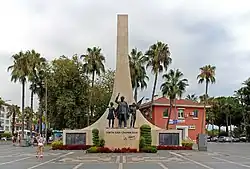 A tall sweeping stone triangle projects skyward behind the statues of a man and two children in bronze on a smaller podium. Around the base are placed several wreaths with logos. Palm trees surround the scene.