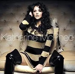 The cover features Katharine McPhee wearing a long black-and-beige shirt and black knee-high boots, sitting against a beige sofa couch. The artist's name appears in front of her, colored in white.