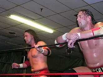 Two white males wearing red wrestling gear standing in a wrestling ring with red and black ropes.