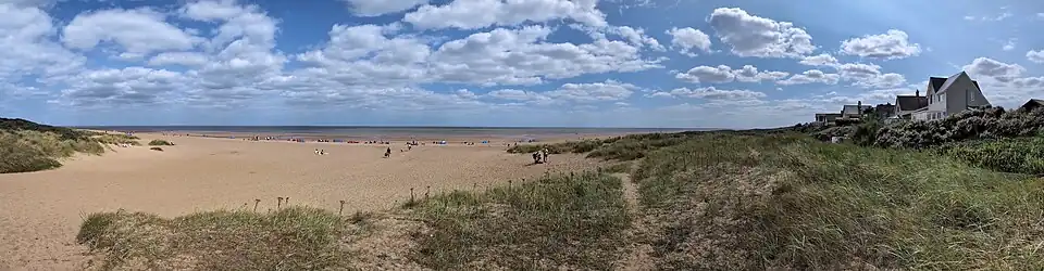 A wide panoramic view of the beach at Anderby Creek, Lincolnshire, England, on a summer weekend. The image shows the expansive sandy foreshore at low tide, looking out towards the North Sea. The beach is backed by sand dunes covered in marram grass, with the houses of the small coastal village visible on the right.