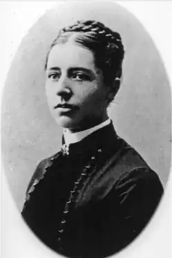 A Black and white portrait photo of a young woman, wearing a black outfit with a high white collar. Her hair is plaited and coiled on the top of her head. Her body is facing to the left of the camera, and she is looking into the camera.