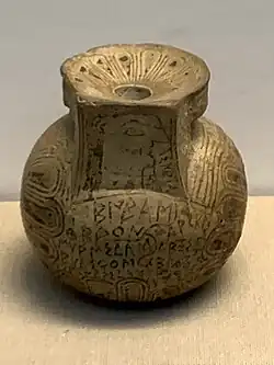 Photograph of a Greek pot, light yellow in colour and covered in writing and drawings, in a museum case.