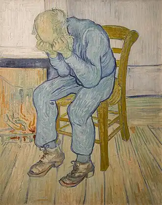 An elderly man with a bald head is sitting on a yellow chair by his fire. There is a low fire in the grate. He is dressed in blue clothes. He is holding his head in his hands.