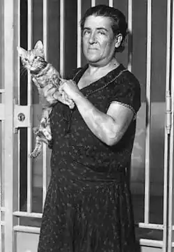 B&W portrait photograph of a standing, middle-aged woman, wearing a dress, holding a cat.