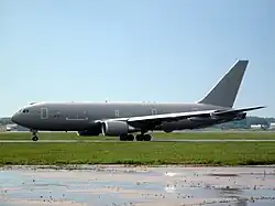 Gray jet aircraft facing left on apron against a cloudless, pale blue sky. In the foreground are green grass; the foreground is a wet tarmac.