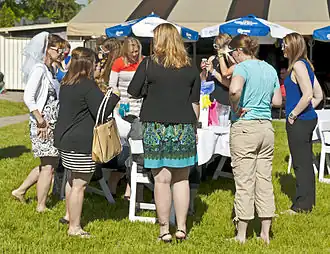 A group of women gathered around a table in an outdoor setting, dressed in a mix of casual and dressy styles. At the left of the image is a woman wearing a veil on the back of her head.