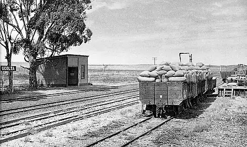 Photo of open railway wagons, a sign reading "Ucolta", and a wooden station building