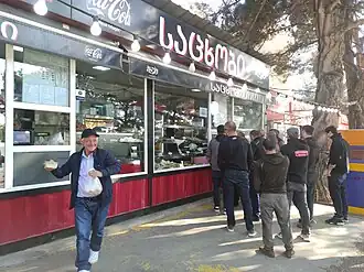 A group of men are gathered at the counter of an outdoor bakery kiosk. A man in the foreground, smiling, walks toward the camera holding a white bag and a piece of cheese.