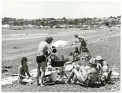 A group of people on a beach sitting around a barbecue grill.