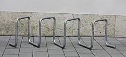 A free-standing bike rack, consisting of galvanized steel tubing bent into multiple square loops set at 45 degrees, on concrete slabs in front of a concrete wall