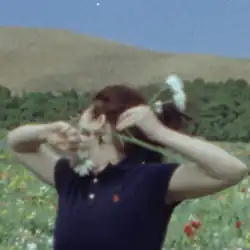 In a grainy image, Eilish in a blue short sleeve polo stands in a meadow of flowers, with a forest and mountain behind her. Her face is turned away from the camera and she is holding a white flower in her arms, raised towards the camera.