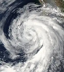 A photograph of a tropical storm off the Pacific coast of Mexico. Most of the thicker clouds are on the right side of the storm, particularly in a long curved band that arcs to the south.