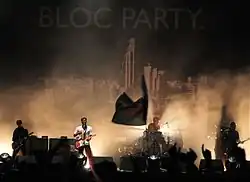Two guitarists, a drummer, and a bassist are performing on a smoky stage lit by white stage lights. The stage background is a black tarpaulin emblazoned with "BLOC PARTY." and a large black-and-white cityscape below it. Most of the crowd have their hands up in the foreground, which also includes a waving flag.
