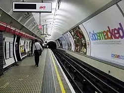The interior of a building with a rounded ceiling and walls and doors on the left, a railway track on the right, and people walking in the middle
