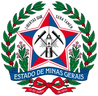 A coat of arms with a large red star in the middle bearing mining equipment. Above the star are the words "Libertas Quae Sera Tamen." The coat of arms is surrounded by a wreath of red berries and green leaves, with two stems of leaves sprouting from the bottom and framing a banner stating "Estado de Minas Gerais." A green ribbon is at the bottom; on the left tail, it says "15 de junho" and on the right, "de 1891."