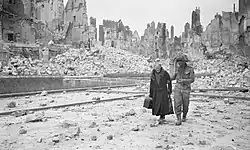 A soldier holding the arm of an elderly lady in a debris-strewn street, with ruined buildings in the background