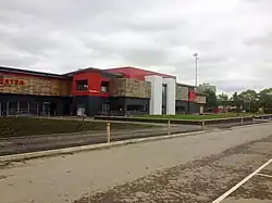 Broadhurst Park football ground on an overcast day, with an access road in the foreground. Visible are two white towers forming the front of the stadium, with a balcony to the left of the left tower. One of the unlit stadium floodlights is visible in the background.