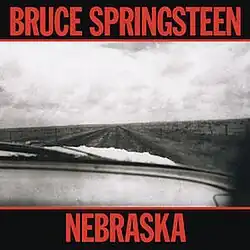 A black-and-white photograph of a desolate landscape taken from inside a car. The words "Bruce Springsteen" and "Nebraska" appear above and below, respectively, in bright red letters.