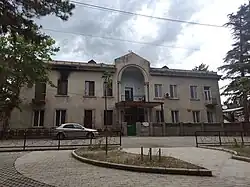 A two-story building with a weathered facade and a prominent arched entrance, located at 3 Samgori II lane in the Tetrikhevi settlement of Tbilisi, Georgia. A noticeable scorch mark from a past fire is visible on the upper left side. The structure shows signs of residential use, including modern windows and an air conditioning unit.