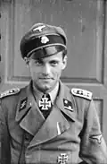 A man wearing a military uniform, peaked cap and a neck order in the shape of a cross. His cap has an emblem in shape of a human skull and crossed bones.