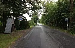 An image of an asphalt road that runs through forests and fields into the distance, with two bus stop signs on both sides of the road.