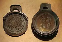 Two circular bronze plates with square handles. The plate on the right is thicker, has four characters embossed into it, and contains a rim around about three-fourths of the edge of the plate. The plate on the left is thinner, and contains a rim that, when the two plates were stacked on one another, would perfectly meet the edges of the first plate.
