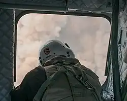 A man in a white Cal Fire helmet and padded clothing cranes out the side of a helicopter door, with only thick smoke visible beyond his head