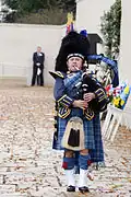 Piper in full Highland Dress with Feather Bonnet in place of Glengarry and blue-grey Doublet with gold braid in place of black with silver braid