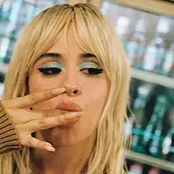 A young woman with long bleached hair, decorative nails, and make-up sticks her middle finger into her mouth as she looks to her left (blurred background of store shelf at a diagonal).