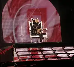 A faraway image of a blond woman sitting on a throne, atop a flight of stairs, on a stage. Two male dancers in black coat and hat, stand in front of her. A red colored backdrop is visible behind the throne.