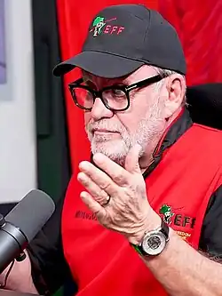 A man seated, white beard, wearing a black baseball cap, black thick-framed glasses, and red polo shirt