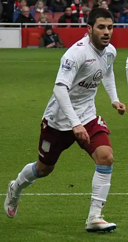 A male soccer player is wearing a white Aston Villa kit and is running.