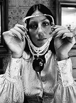 Cher holds up an ornate multi-strand beaded necklace with a large pendant, wearing a paisley-patterned blouse with billowy sleeves.