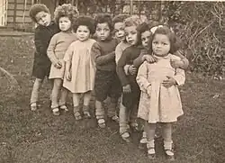 8 mixed race children standing in a row in a garden in the 1940s at Holnicote House in England. The children are wearing dresses or shorts.
