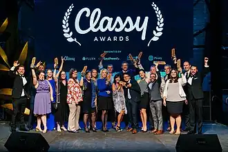 A photograph of twenty people standing in two rows on a stage. They are nicely dressed and holding up award statuettes. The lighting is very blue and behind them is a screen with the Classy logo and sponsors projected.