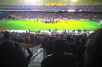 A team celebrates in the center of a soccer field while fans in stand on both sides cheer.