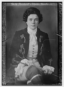 A young white woman dressed in an opera costume, embroidered jacket and 18th-century wig, seated in a chair with legs crossed