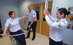 A test person is sitting in a swivel chair. A second person has just stopped the rotation of the chair. The disoriented test person points at a sign held up by a third person.