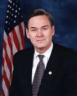 A gray-haired man in a suit sits in front of an American flag