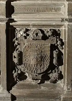 This image shows a detailed view of the carved tomb chest from the monument to Sir Christopher Hildyard at Winestead, Yorkshire East Riding. The inscription panel at the top records his death date of 1530. Below is an elaborately carved heraldic achievement featuring the Hildyard coat of arms as the centrepiece, surrounded by ornate Renaissance-style decorative elements. The carving demonstrates the transition from Gothic to Renaissance ornamentation typical of early Tudor monuments, with flowing foliate scrollwork, grotesque masks, and classical motifs framing the heraldic shield. The quality of the stonework is exceptionally fine, with deep undercut carving creating dramatic shadows and three-dimensional effects. Such heraldic displays were crucial elements of aristocratic monuments, proclaiming family lineage, alliances, and social status. The combination of heraldry with Renaissance decorative vocabulary shows how English monument design was beginning to incorporate Continental influences whilst maintaining traditional English emphasis on armorial display during the early sixteenth century.