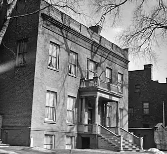 A black-and-white photograph showing a brick house from its front right. There is some remaining snow on the ground, and the top is partially obstructed by bare tree branches. The front of the house has an ornate portico with columns and a flat roof.